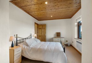 A bedroom with a wooden ceiling, a bed, a nightstand, a dresser, and a window. - Rugosa Family House in Scenic Ballyconneely (Ballyconneely)