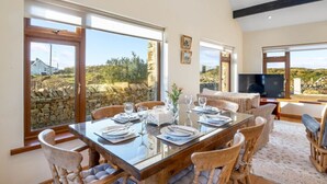 Un comedor con una mesa grande preparada para seis personas, ventanas amplias con vistas a un muro de piedra y vegetación, y un televisor de pantalla plana al fondo.