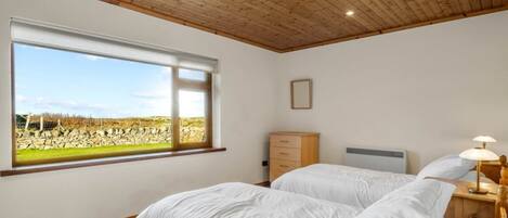 A bedroom with two beds, a large window with a view of a field, wooden ceiling, and a nightstand with a lamp.