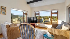 Amplia sala de estar con muebles de mimbre, televisor de pantalla plana y ventanas grandes que ofrecen vistas al campo.