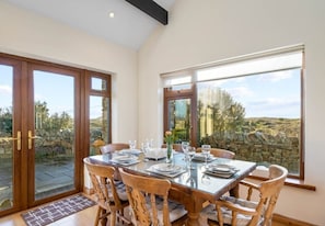 A dining area with a table set for four, large windows, and a stone wall outside. - Rugosa Family House in Scenic Ballyconneely (Ballyconneely)