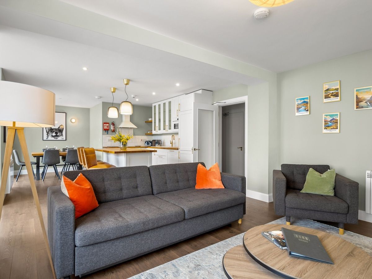 A modern living room with a grey sofa, orange and green cushions, a coffee table, and a dining area in the background.