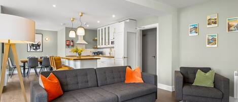A modern living room with a grey sofa, orange and green cushions, a coffee table, and a dining area in the background.
