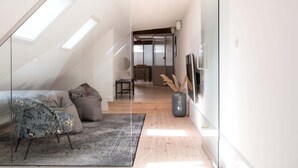 A modern loft bedroom with a sloped ceiling, skylight, a large window, a bed, a sofa, a rug, and a TV. - Nyhavn63 Suites 2 by Daniel&Jacob's (København)