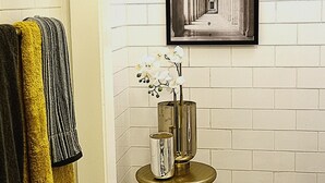 Ett badrum med ett rutmönstrat golv, en inramad tavla på väggen och en vas med blommor på ett piedestal.