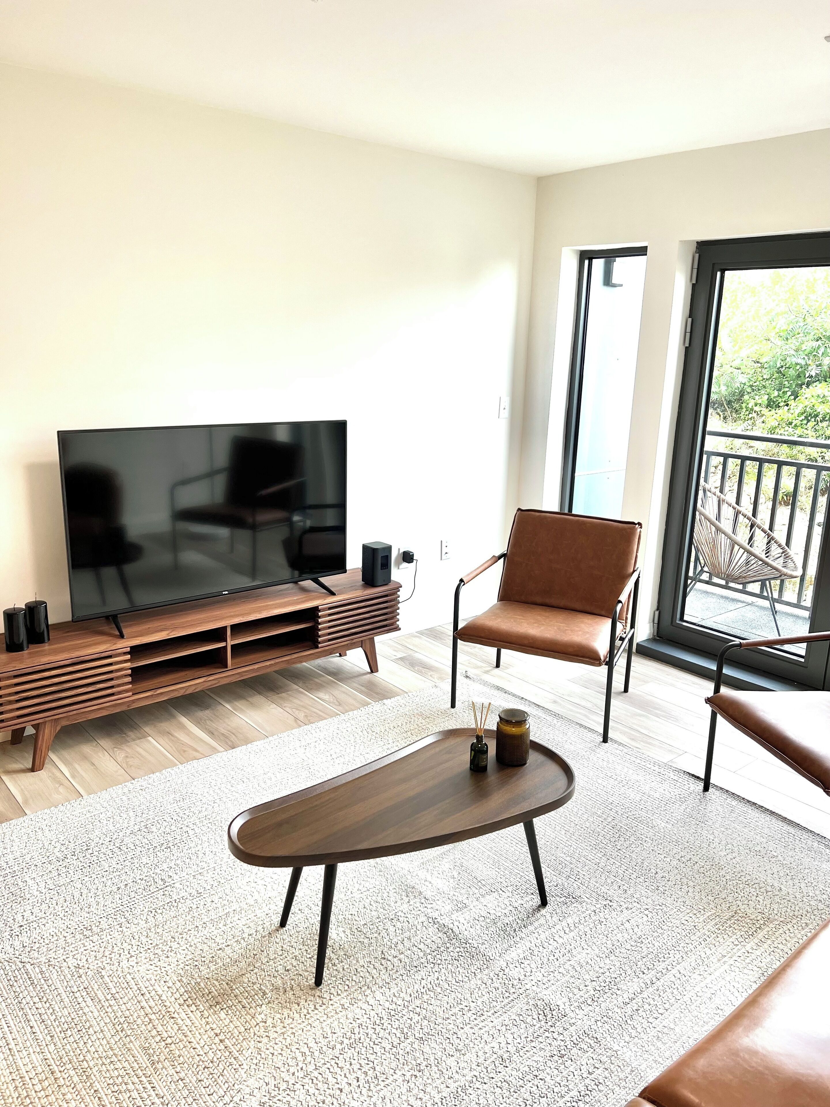 Una sala de estar moderna con un televisor de pantalla plana sobre un soporte de madera, un sillón de cuero marrón, una mesa de centro redonda y un ventanal con vistas a zonas verdes.