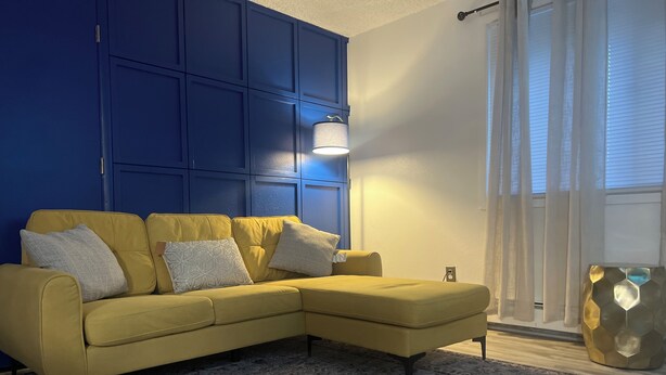 Ein gelbes Ecksofa mit gemusterten Kissen, eine blaue Akzentwand, ein Teppich mit Muster und ein großes Fenster mit transparenten Vorhängen.