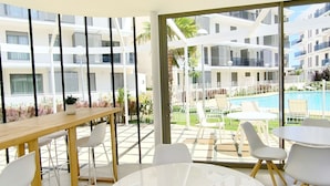 A modern hotel room with a large window, a wooden table, white chairs, and a view of a pool and other buildings. - Ático Marina Real III (Dénia)