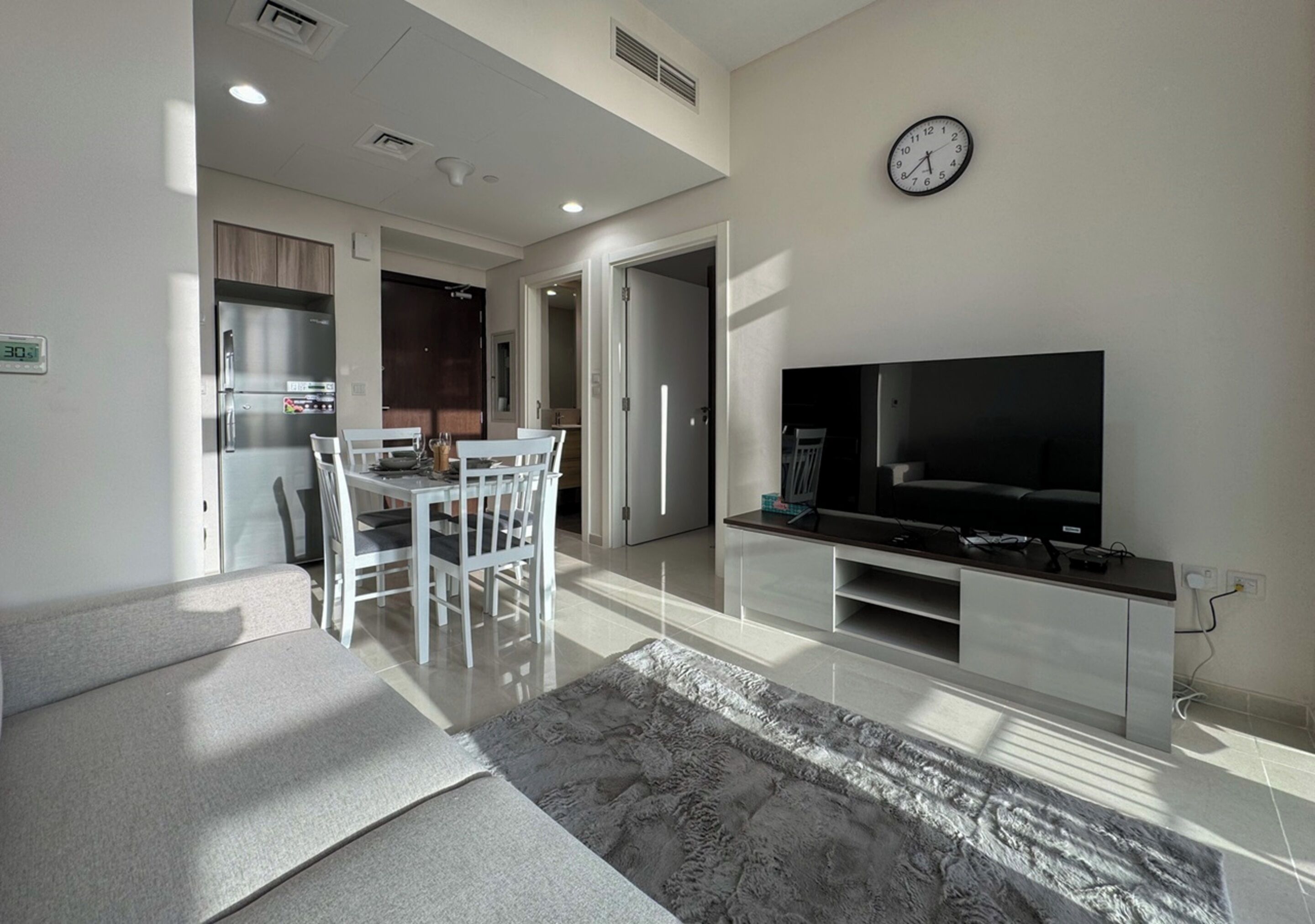 A modern living room with a flat-screen TV, a dining area with white furniture, and a kitchen in the background.