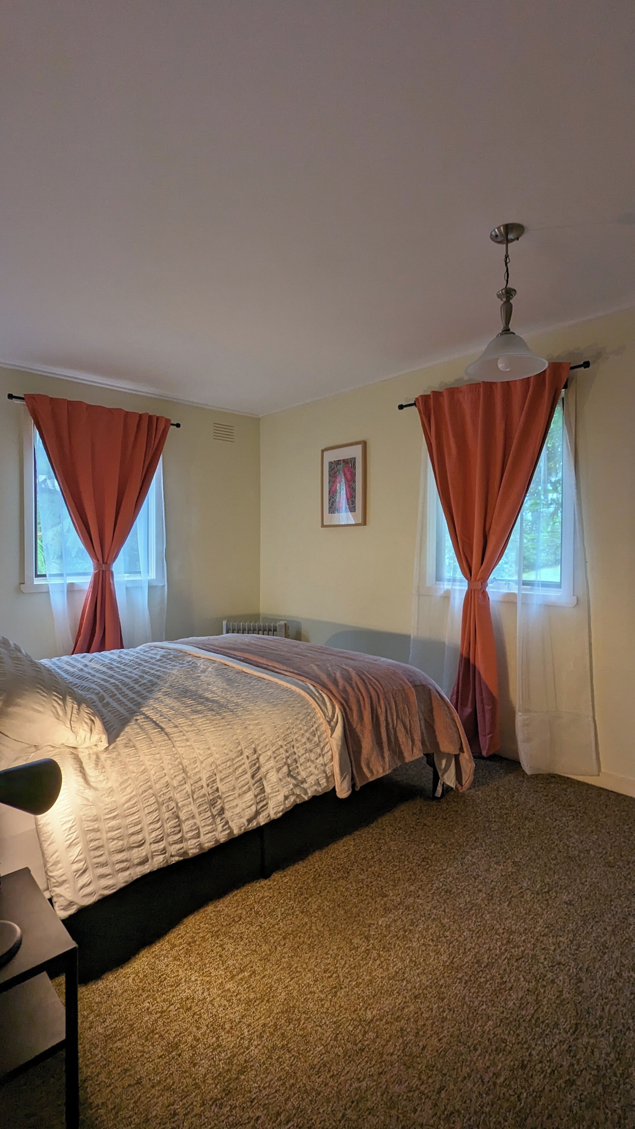 A bedroom with a bed, two windows with curtains, a radiator, and a ceiling fan.