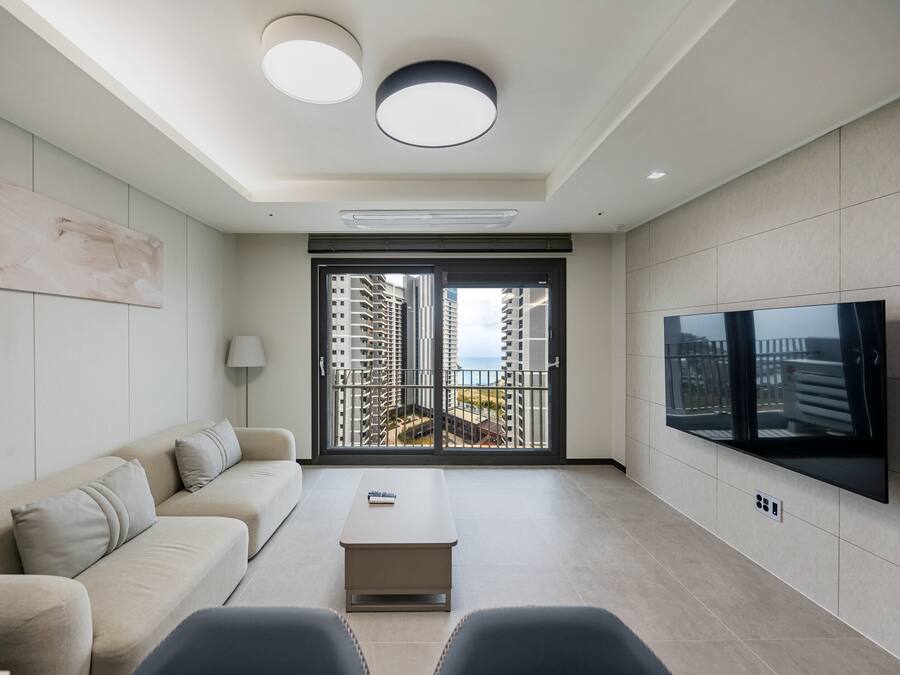A modern living room with a sofa, a coffee table, a flat-screen TV mounted on the wall, and a large sliding glass door leading to a balcony with a city view.