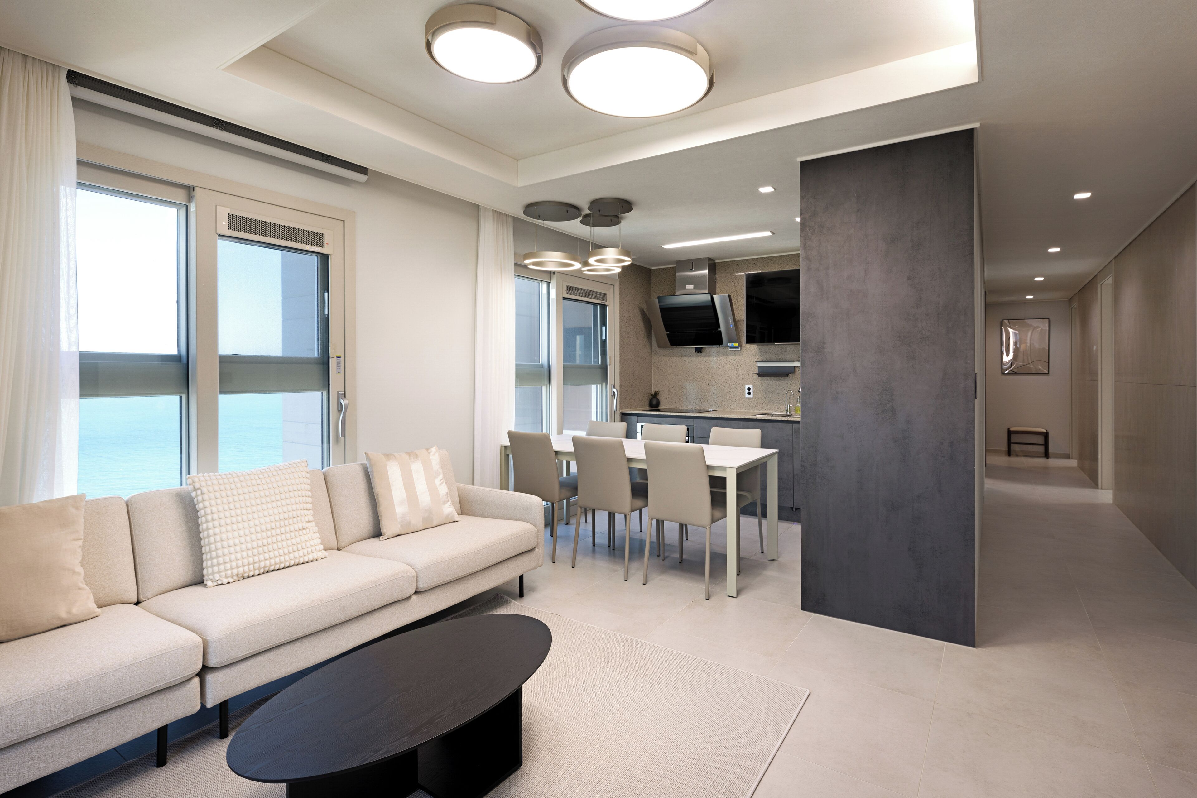 A modern living room with a sofa, dining table, and chairs, a kitchen area, and large windows with curtains.