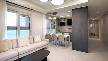 A modern living room with a sofa, dining table, and chairs, a kitchen area, and large windows with curtains.