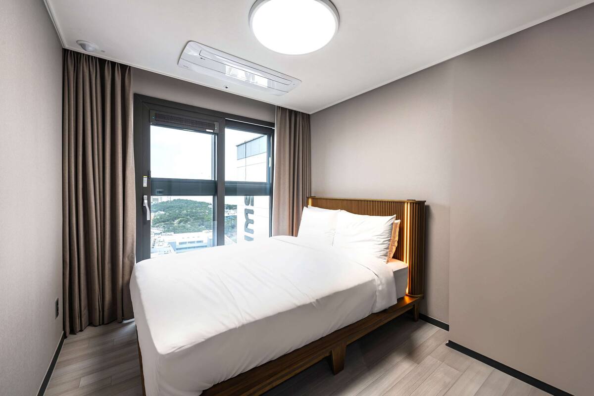 Habitación de hotel con cama, ventana con cortinas, lámpara en el techo y vistas a edificios.