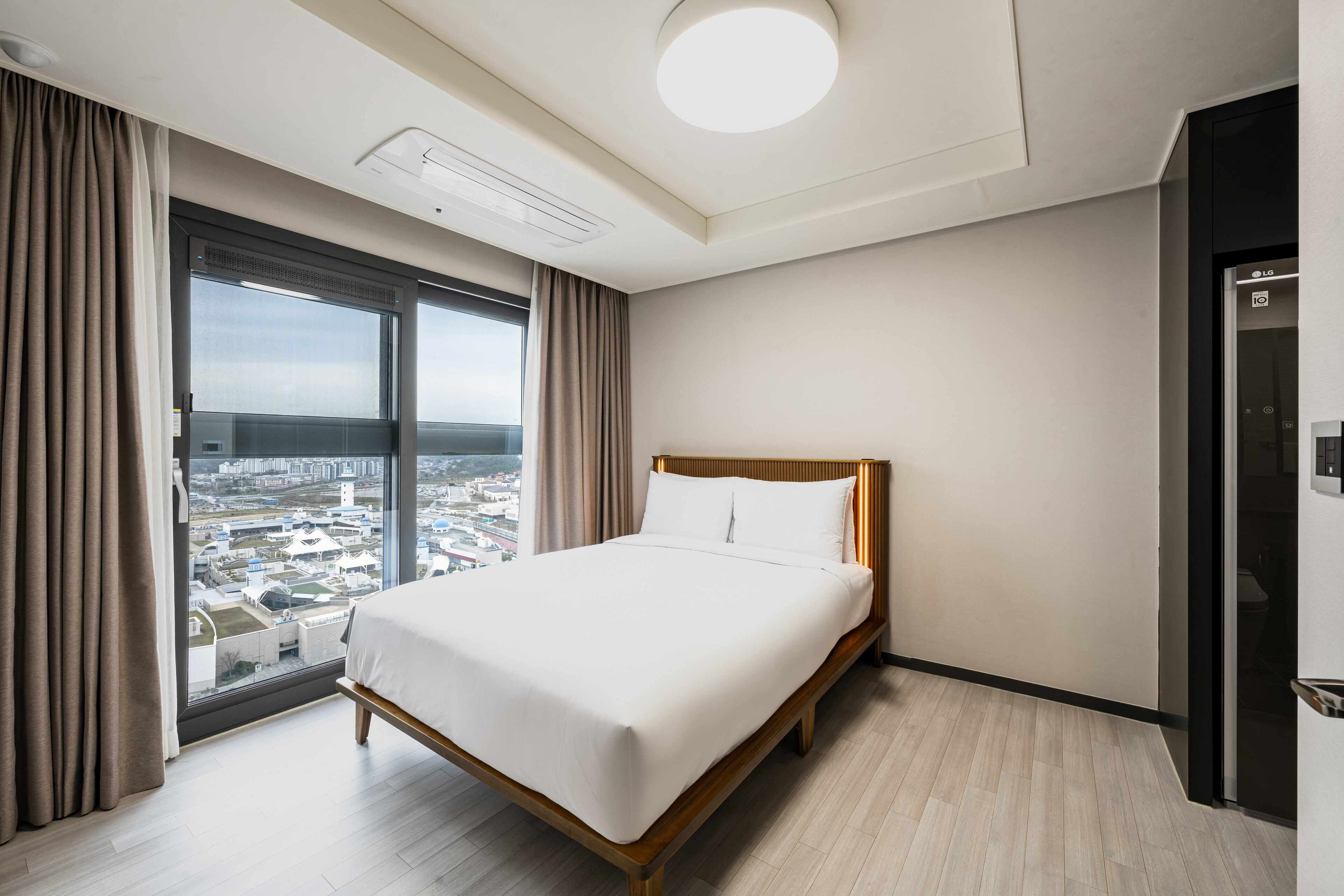 Een hotelkamer met een groot bed, een raam met gordijnen, uitzicht op een stadsgezicht en een moderne plafondlamp.