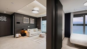 A modern hotel room with a sofa, a small table, a reading lamp, and a large window offering a view of the water. - Odays Osiria Residence (Busan)