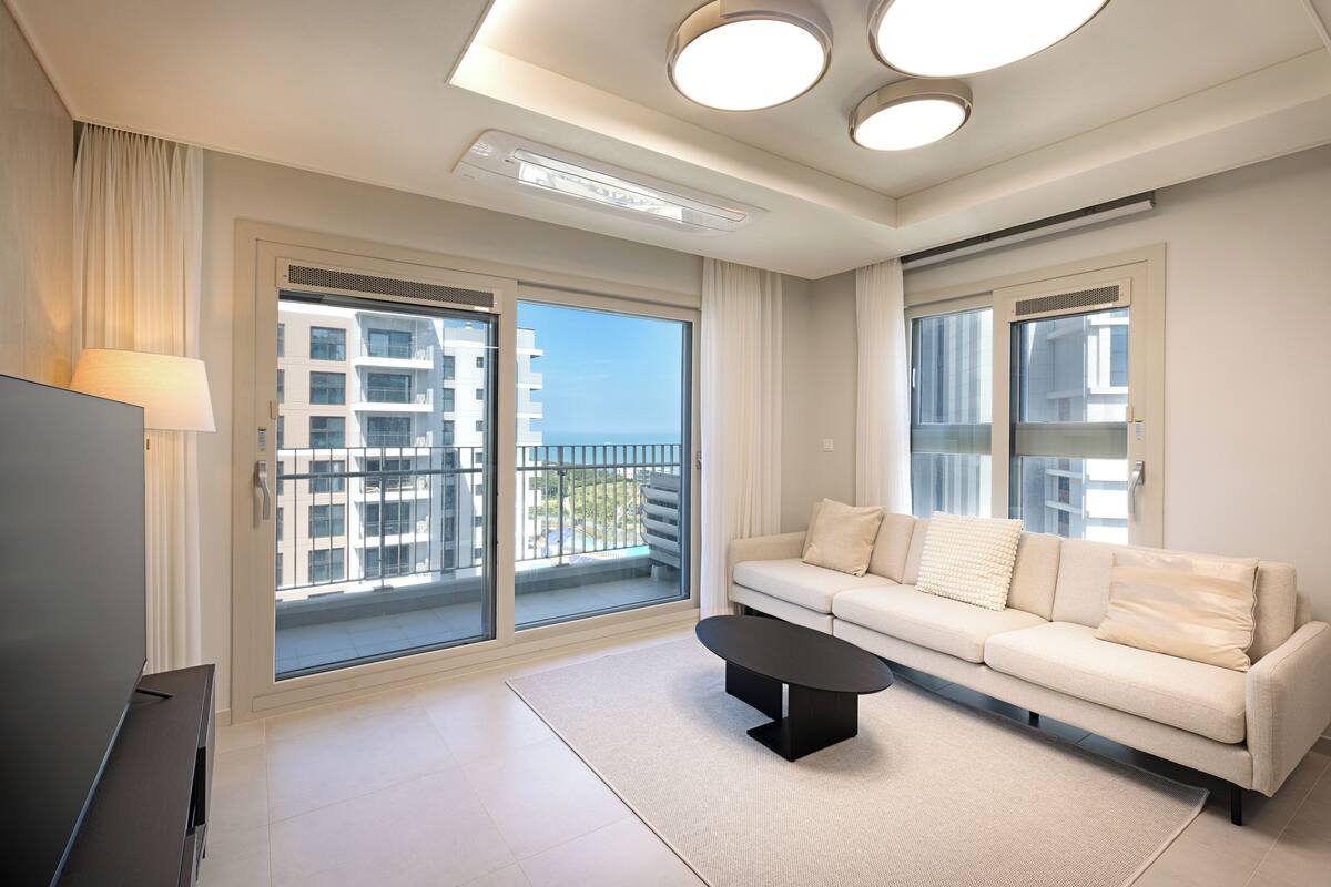 Una sala de estar moderna con un sofá, una mesa de centro y un televisor. Amplias ventanas ofrecen vistas a edificios y un balcón.