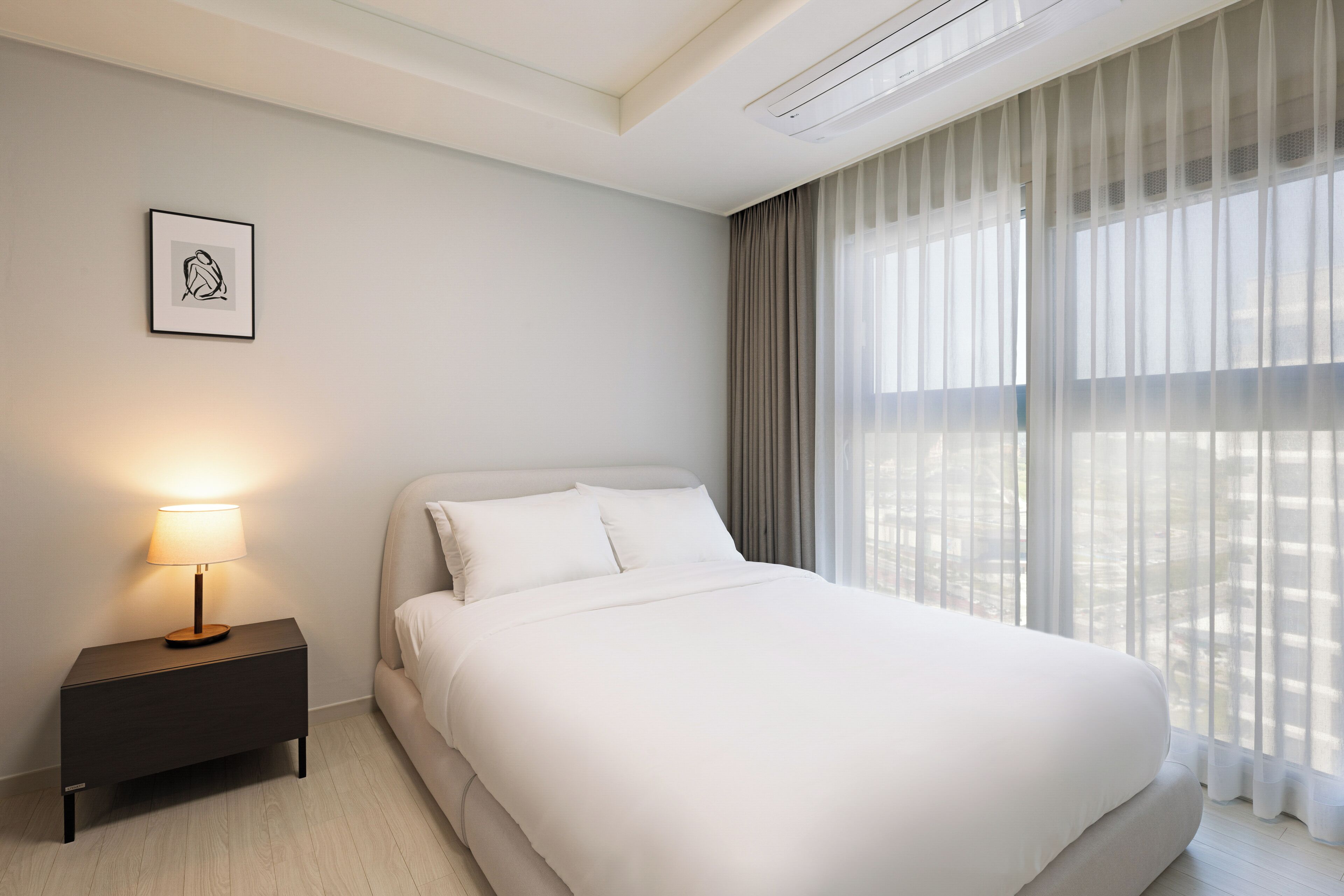 Ein modernes Hotelzimmer mit einem ordentlich bezogenem Bett, einer Nachttischlampe, einem gerahmten Kunstwerk an der Wand und einem großen Fenster mit transparenten Vorhängen.