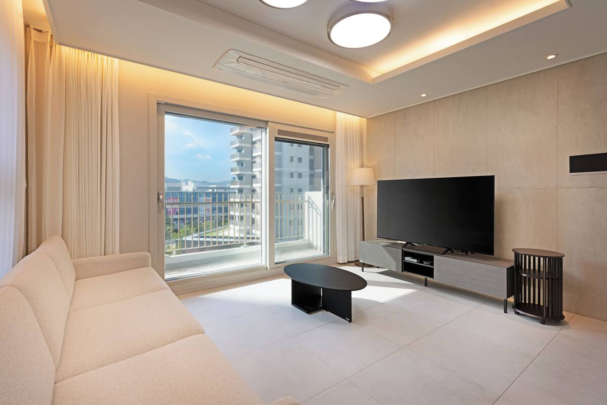 Una sala de estar moderna con sofá, mesa de centro, televisor y un balcón con vistas a edificios.