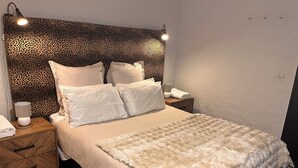 A bed with a leopard print headboard, white pillows, and a fluffy white throw blanket. There is a bedside table with a lamp and a folded towel.