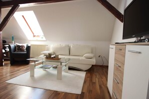 Un espace de vie de style « loft » avec un canapé blanc, une table basse en verre et un téléviseur à écran plat posé sur un meuble en bois.