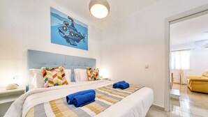 Habitación de hotel con una cama grande, cabecera, un cuadro en la pared y vista hacia otra habitación.