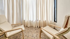 Una habitación con dos sillas color beige, un ventanal con cortinas translúcidas y un suelo de baldosas de piedra.