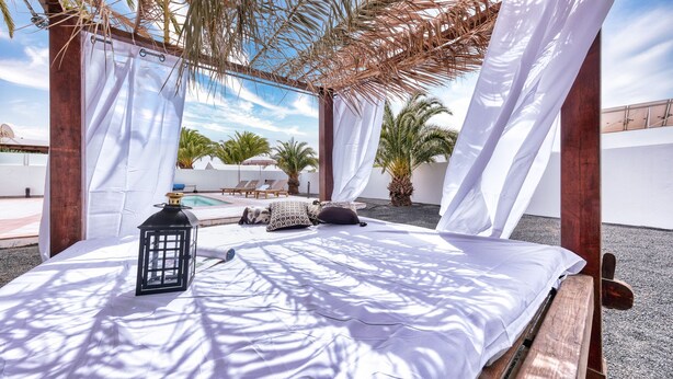 A sunlit outdoor lounge area with a wooden bench, a white canopy, and a pool in the background.