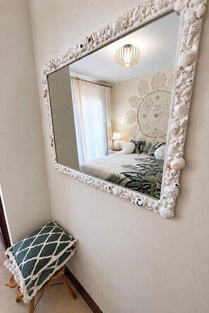 A bedroom with a large mirror, a bed with a floral bedspread, a bedside lamp, and a small stool. - Casa Bleu - Duplex - Pool and BBQ By LVH (Puerto Calero)