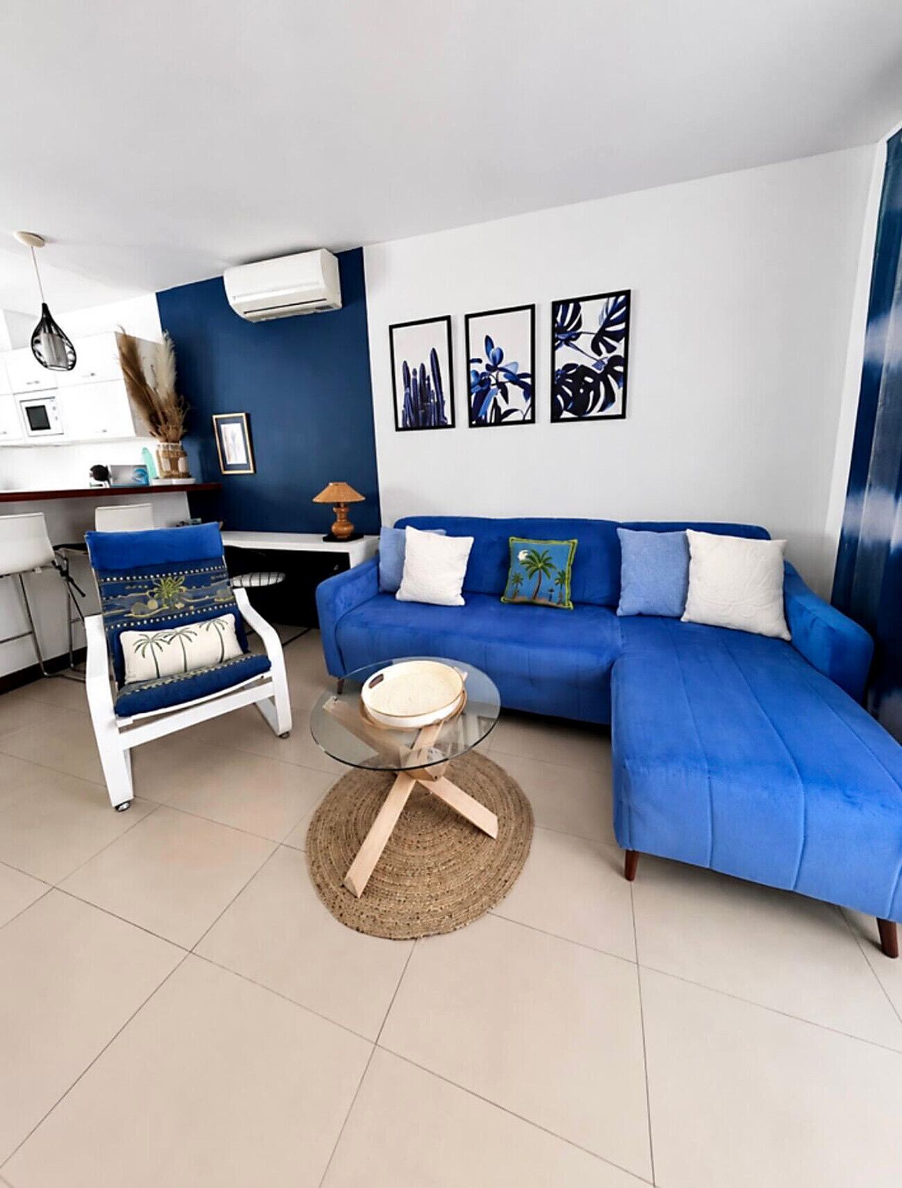 Una sala de estar con un sofá azul, un sillón blanco y una mesa de centro de cristal.