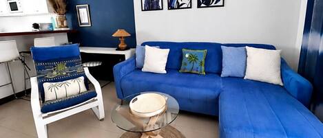 Una sala de estar con un sofá azul, un sillón blanco y una mesa de centro de cristal.