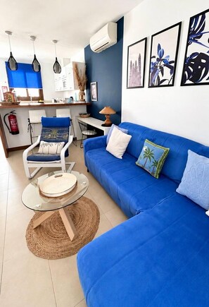A living room with a blue sofa, a round glass coffee table, a white chair with a blue cushion, and a small round rug. - Casa Bleu - Duplex - Pool and BBQ By LVH (Puerto Calero)