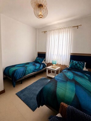 Ein Hotelzimmer mit zwei Einzelbetten, einem Nachttisch mit Lampe, einem Fenster mit Vorhängen und einer besonderen Deckenleuchte.
