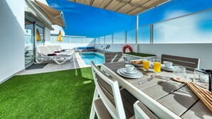 A modern outdoor dining area with a pool, white furniture, and a clear blue sky.