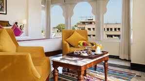 Une chambre d’hôtel avec un lit, deux fauteuils jaunes, une table basse en marbre et une vue sur les bâtiments à travers des fenêtres en arc de cercle.