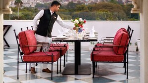 Un homme prépare une table avec des chaises rouges et une nappe blanche, donnant sur un paysage urbain avec des collines.