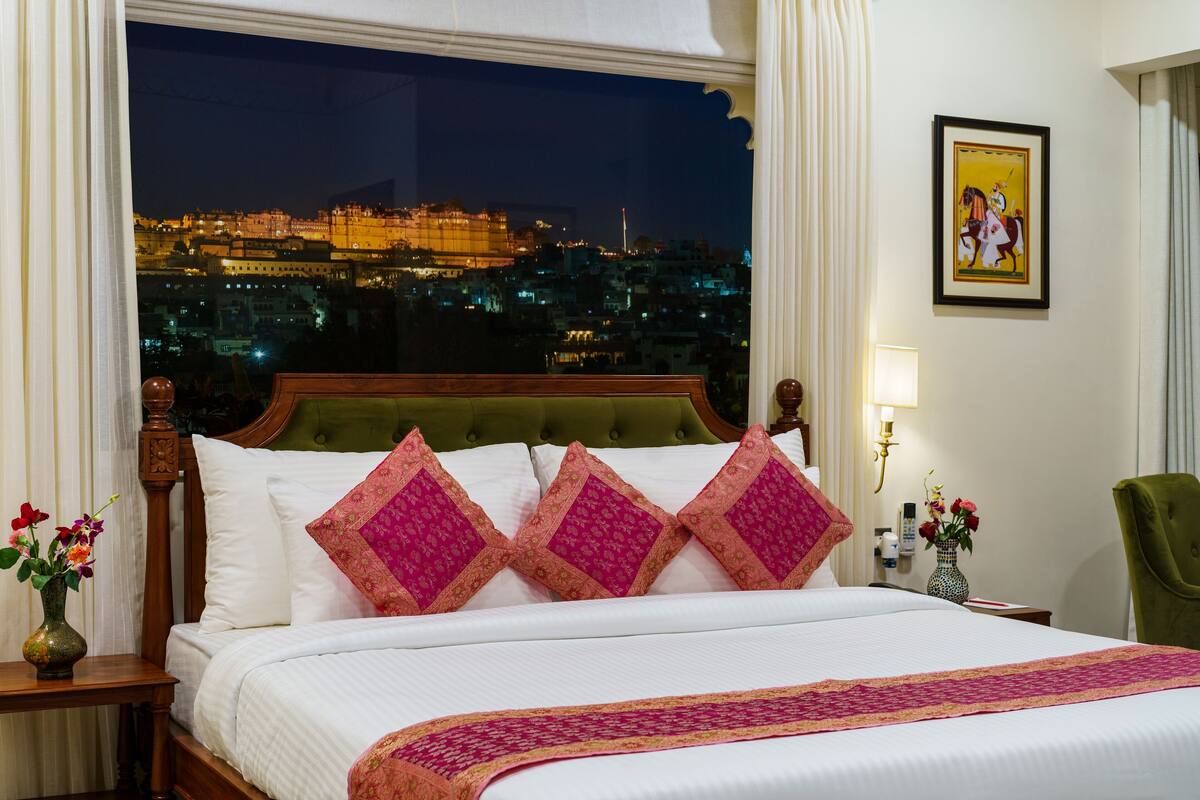Une chambre d’hôtel avec un grand lit, une vue sur le paysage urbain illuminé la nuit, une tête de lit recouverte d’un tissu vert et un tableau encadré au mur.