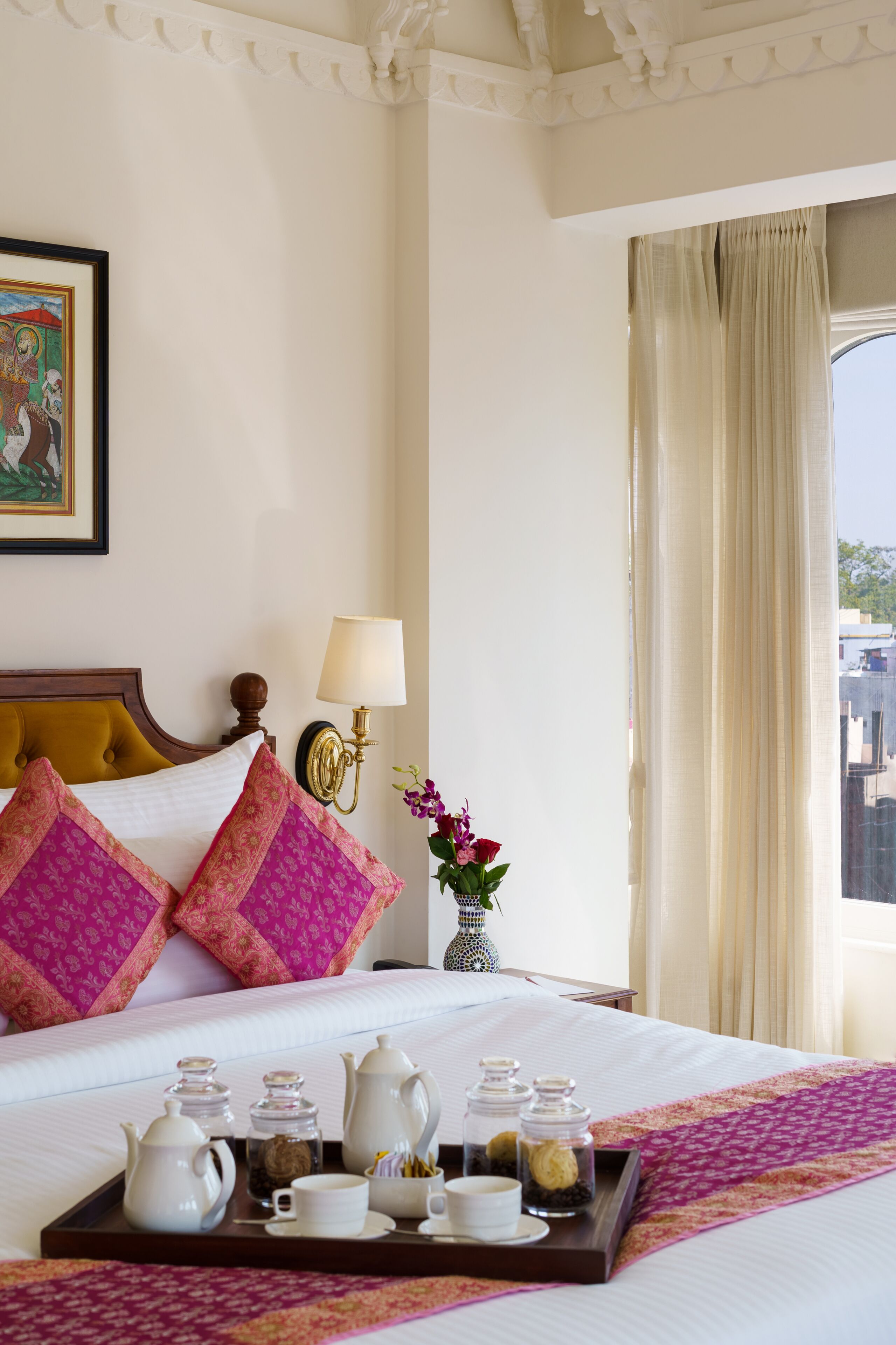 A neatly made bed with a tray of tea and sugar, a vase with flowers, and a painting on the wall.