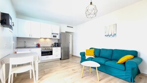 A modern kitchen with white cabinets, a dining table with chairs, a teal sofa with yellow pillows, and a geometric pendant light.
