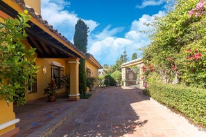 A paved pathway leading to a yellow building with a tiled roof, surrounded by lush greenery and flowering plants.