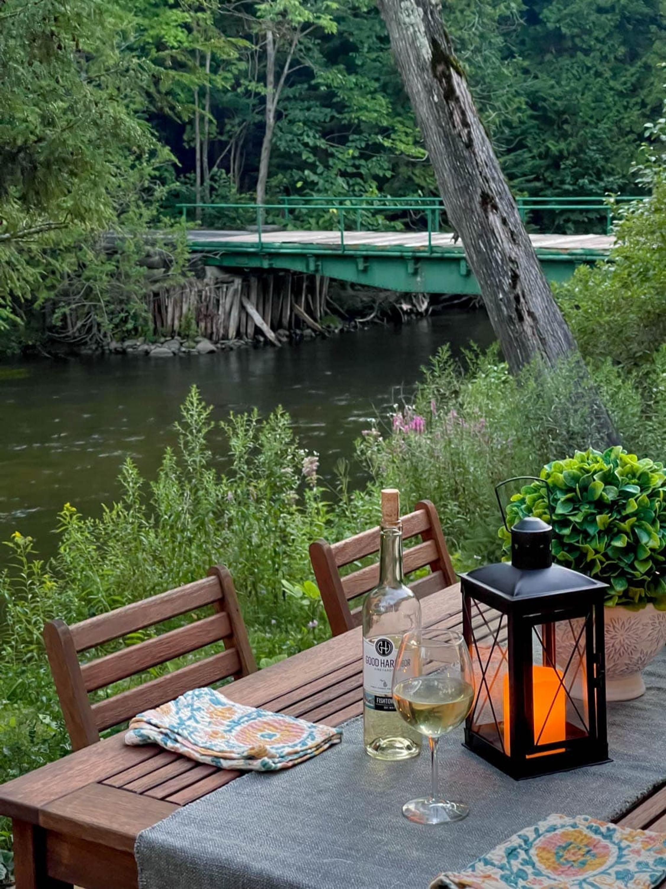 Una mesa de madera con una botella, un vaso, un farol y vistas de un río con un puente.