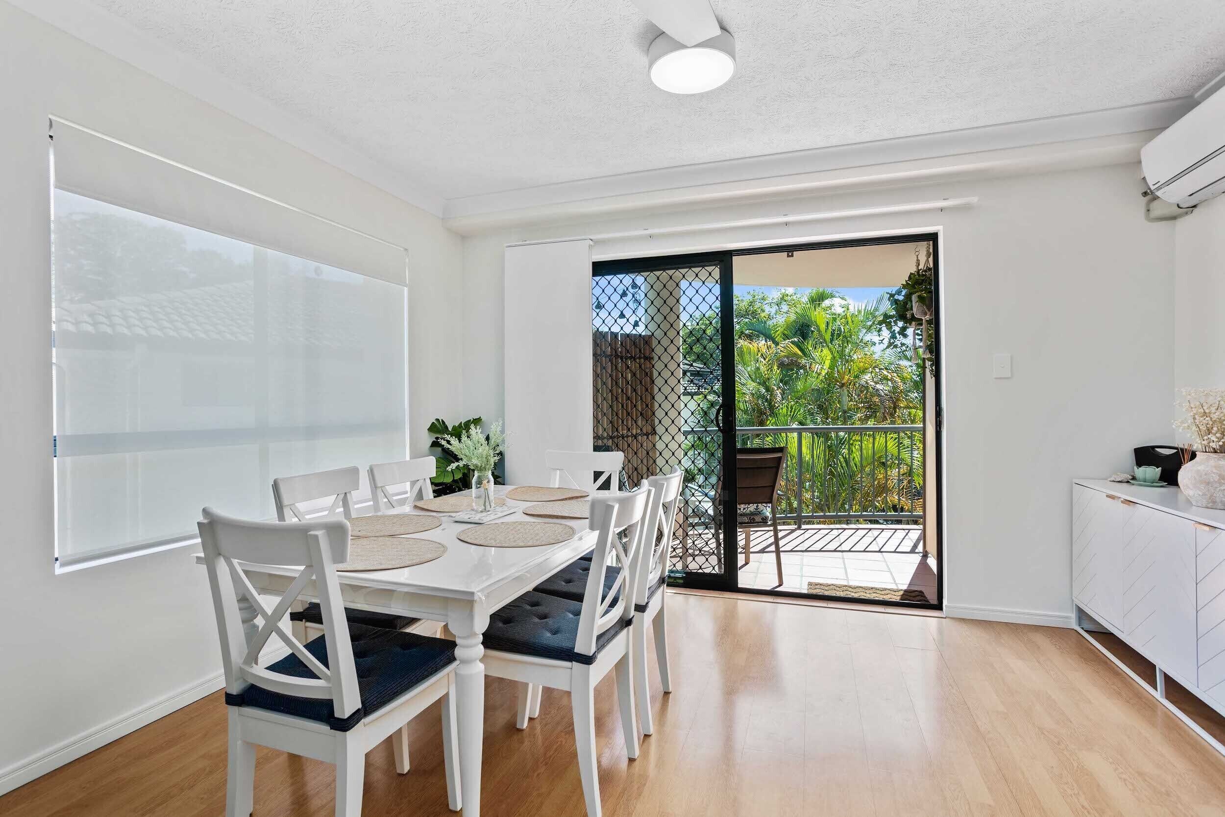 A dining area with a white table and chairs, a white cabinet, and a sliding glass door leading to a balcony with plants.