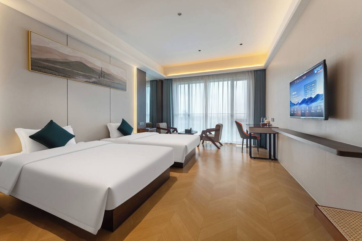A hotel room with two beds, a desk, a chair, a television, and a large window with curtains.