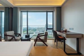 A hotel room with a large bed, a desk, two chairs, and a view of the cityscape through large windows.