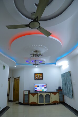 A room with a ceiling fan, recessed lighting, a television on a cabinet, and a door leading to another room. - NANDA MOHAN HOMESTAY (Tirupati)