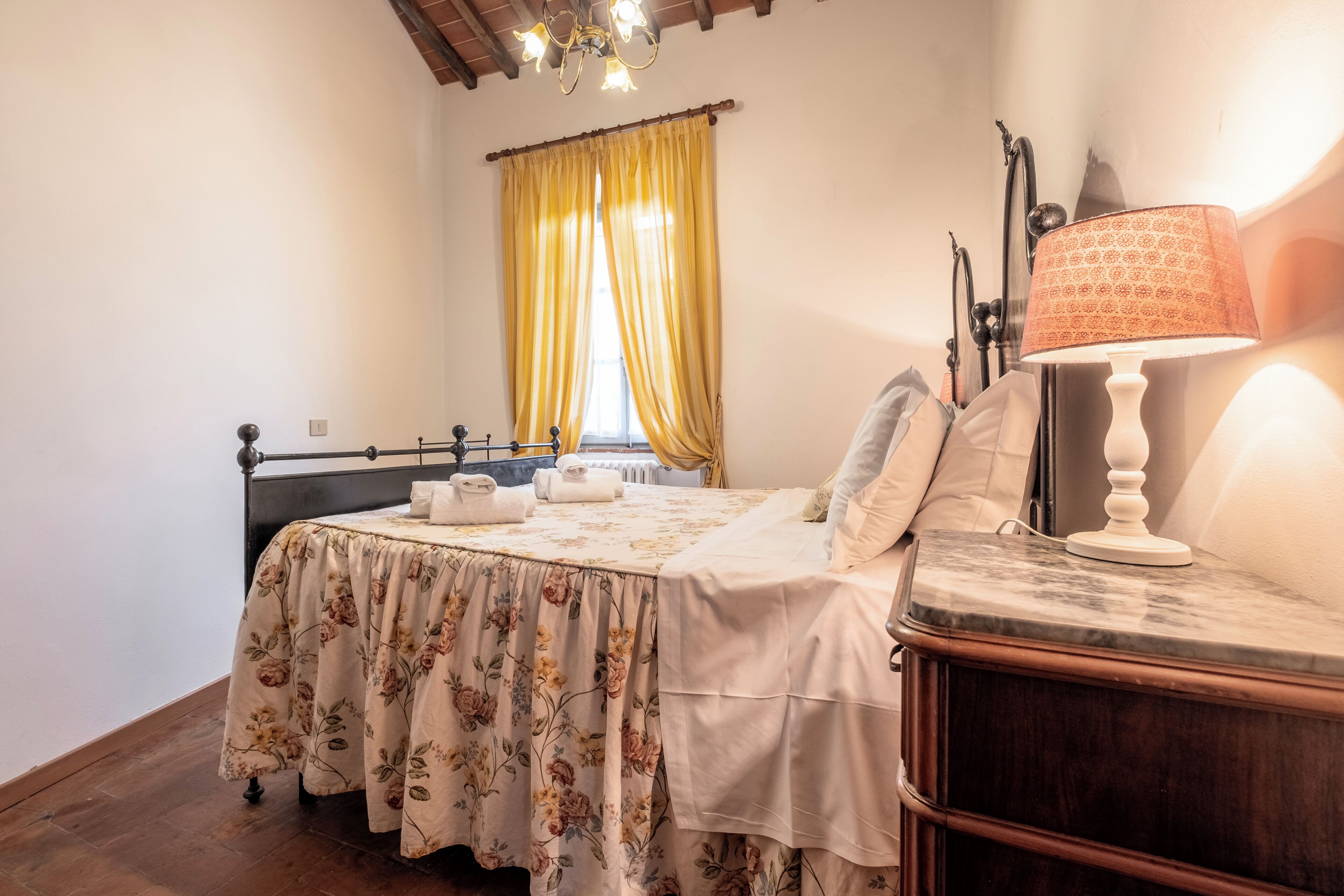 Un dormitorio con cama, mesita de noche, lámpara, ventana con cortinas y techo de madera.