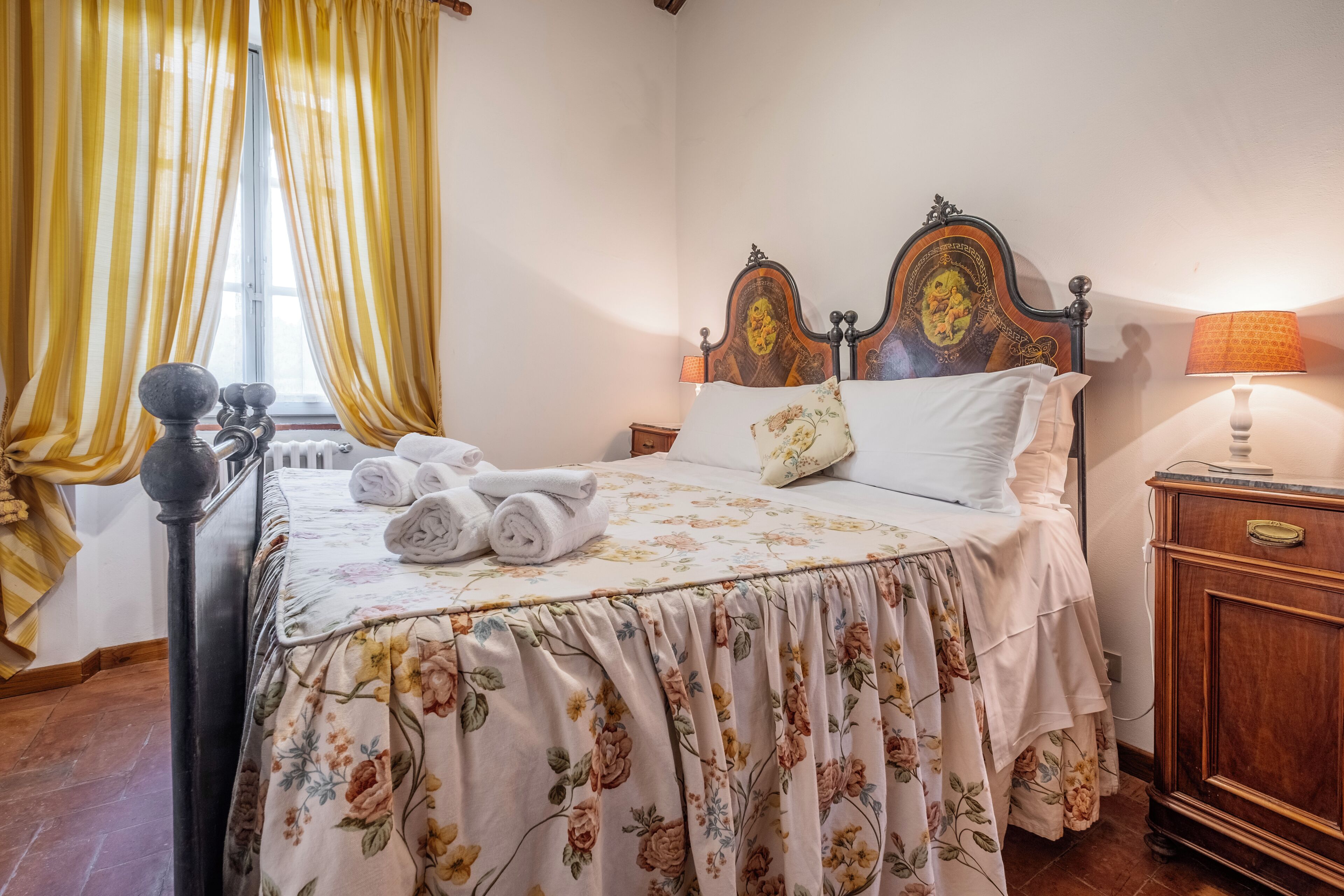 Een opgemaakt bed met bloemenbeddels, een houten hoofdbord en een nachtkastje met een lamp.