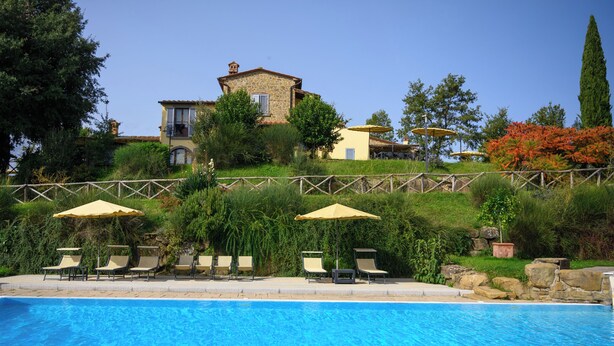 Une piscine avec des chaises longues et des parasols, une maison en pierre avec un jardin et un ciel bleu azur.