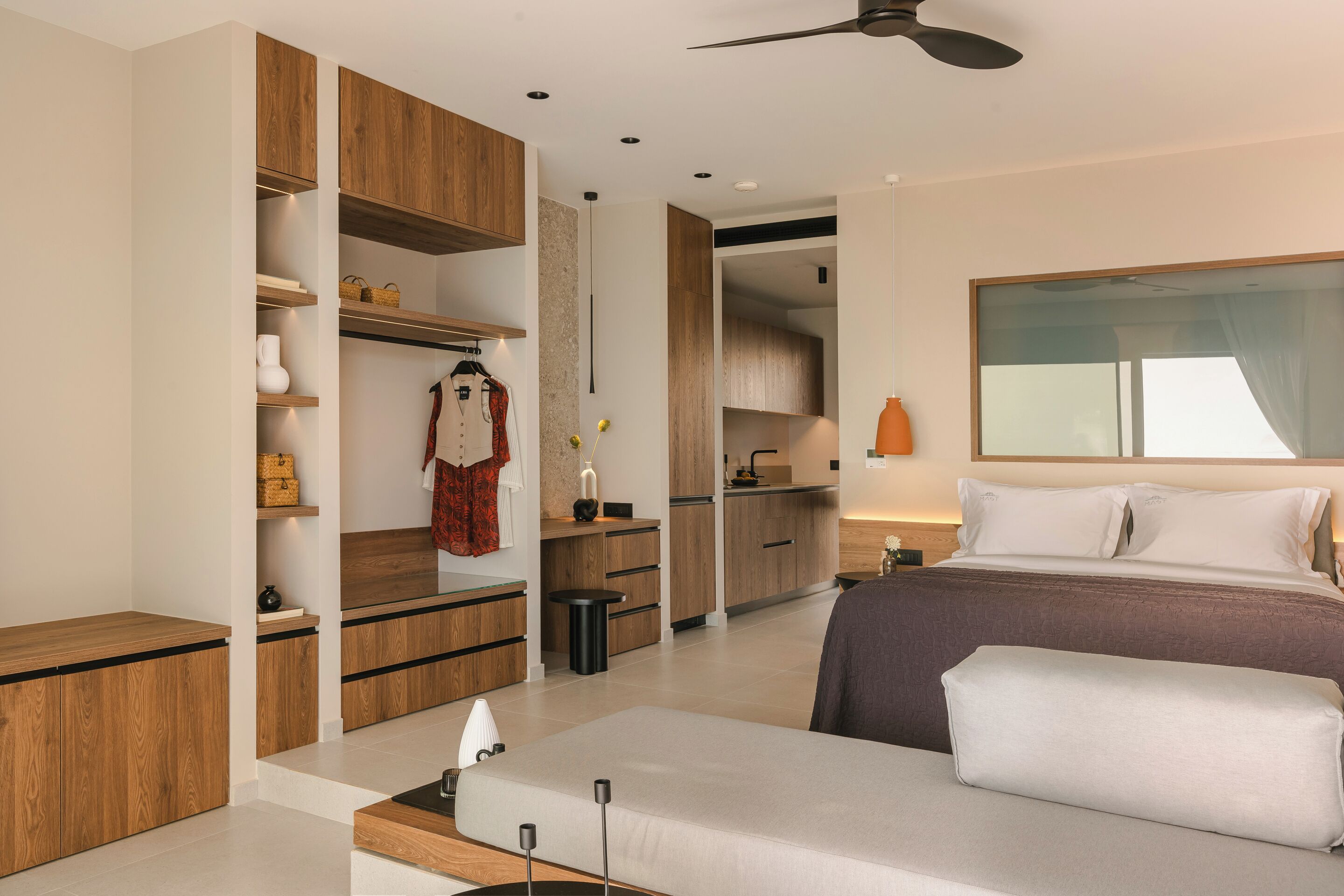 A modern hotel room with a bed, bedside table, wardrobe, and a kitchenette.