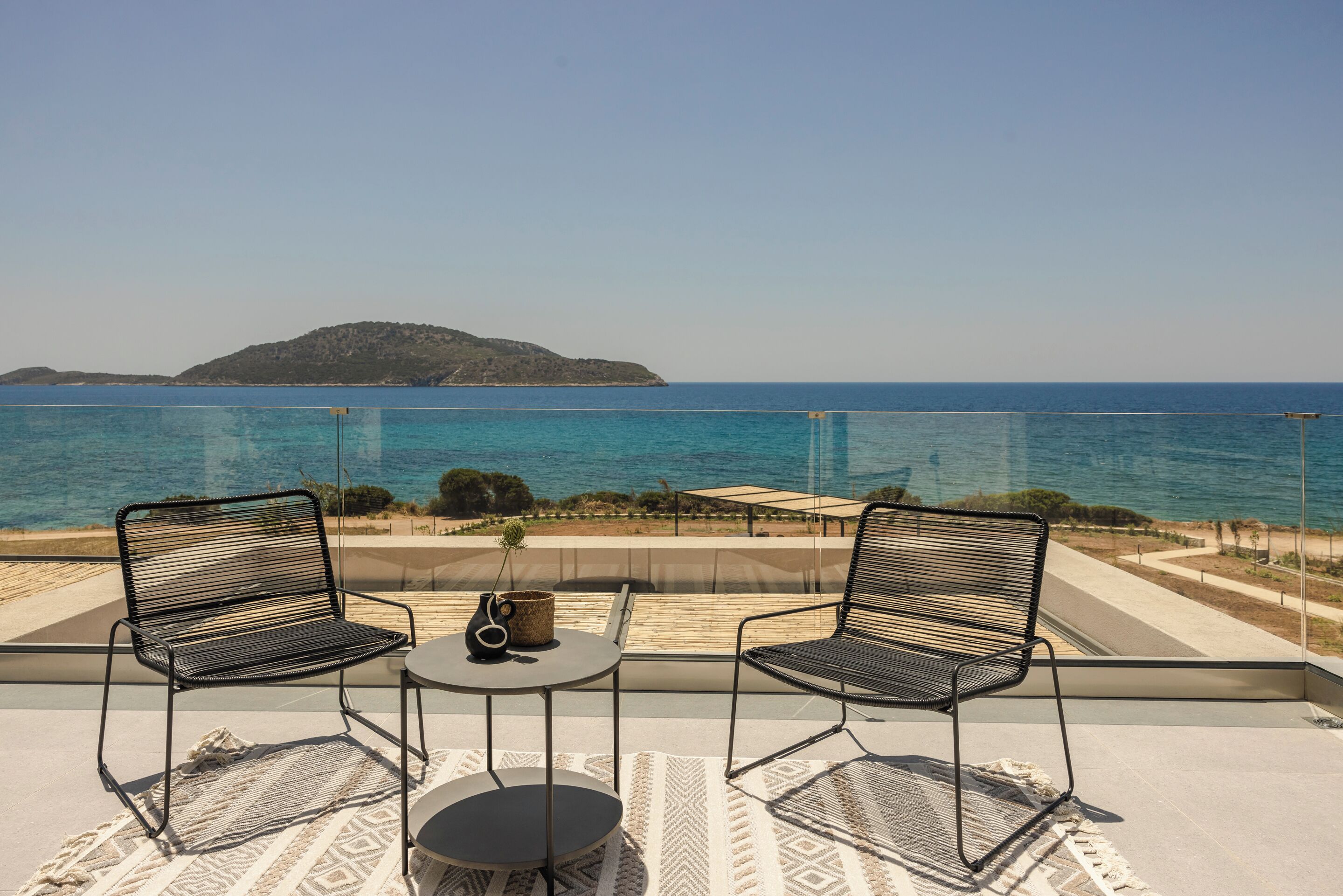 Un balcon avec deux chaises et une petite table, d’où l’on peut admirer une mer bleue et limpide et une île lointaine.